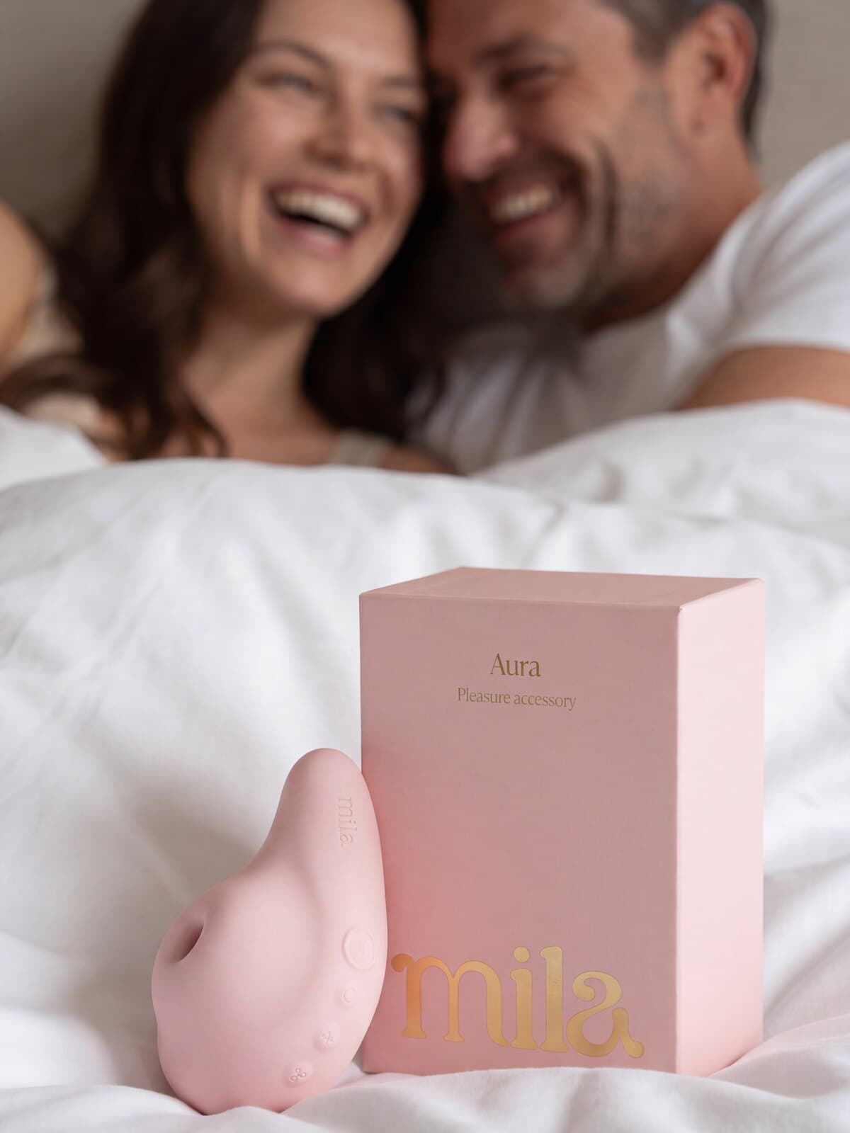 A couple laughing together in bed, the pink Aura massager and its Mila box on the covers in front of them.