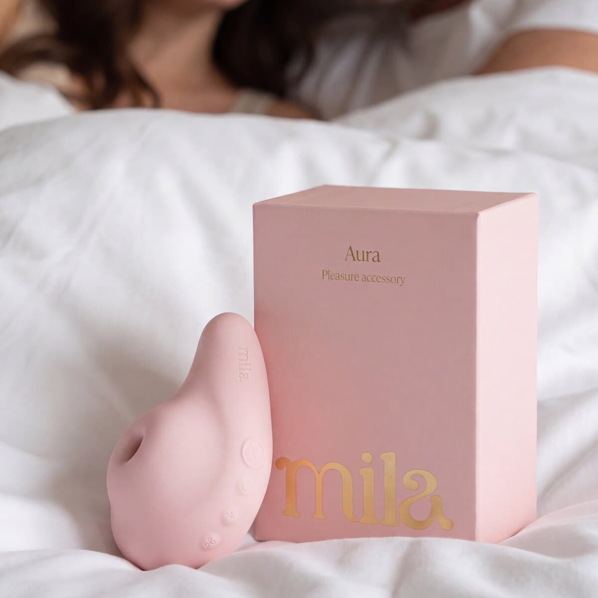 The pink Aura massager beside its gold-lettered Mila box on white bedding, someone resting out of focus behind.