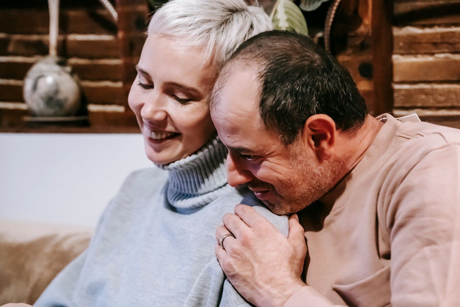 A couple in their fifties sit close on a sofa, her head tipped back against him, both smiling with their eyes closed.