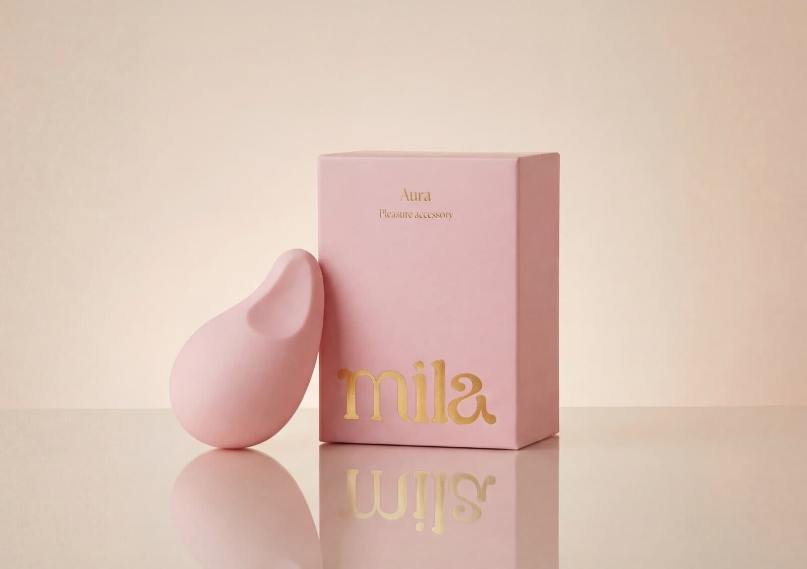 The pink Aura massager leaning against its gold-lettered Mila box on a reflective cream surface.