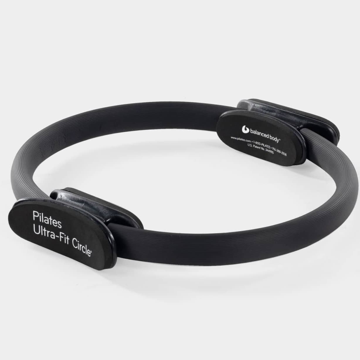 A black Balanced Body Pilates Ultra-Fit Circle with padded handles on either side, photographed on a pale grey ground.