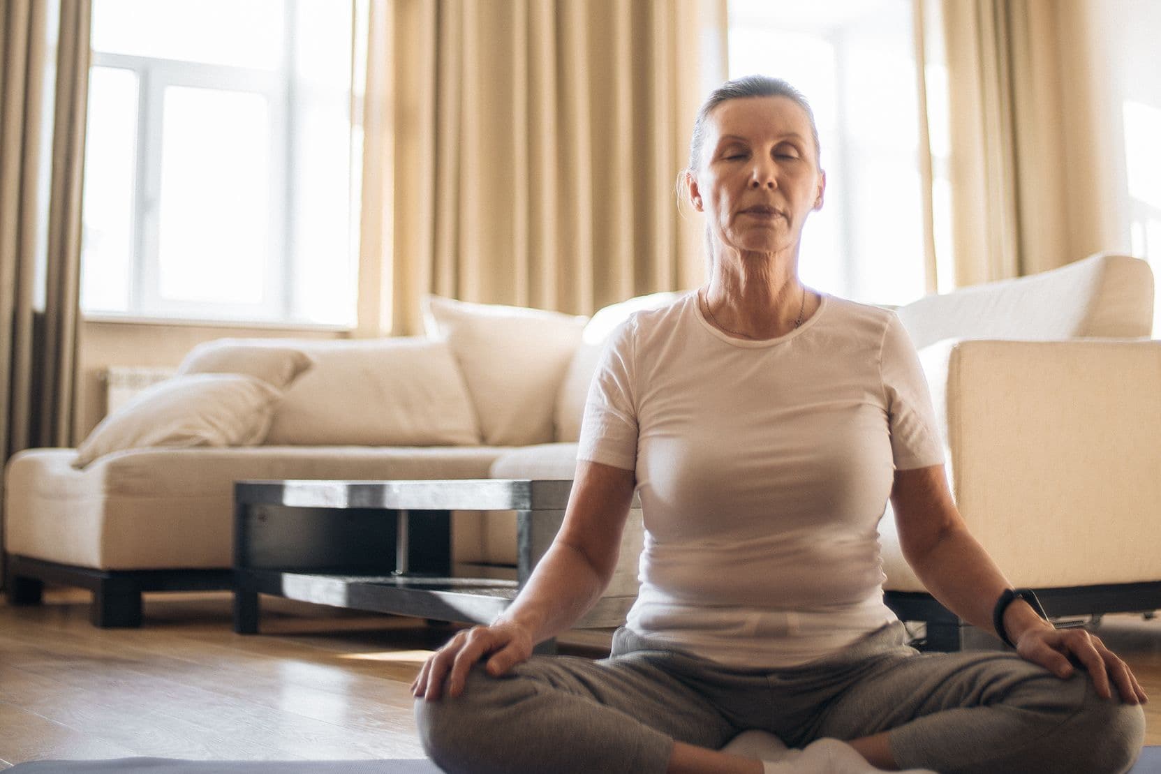 A woman in her sixties sits cross-legged on a mat in a sunlit living room, eyes closed, hands resting on her knees.